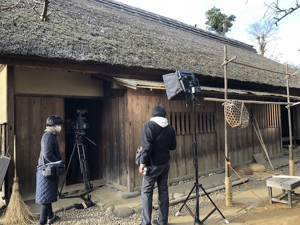 藁葺き屋根の古い平屋の建物の入り口にカメラや照明が瀬地され、竹で作られた物干し竿の横に2名のスタッフがいる撮影風景の写真