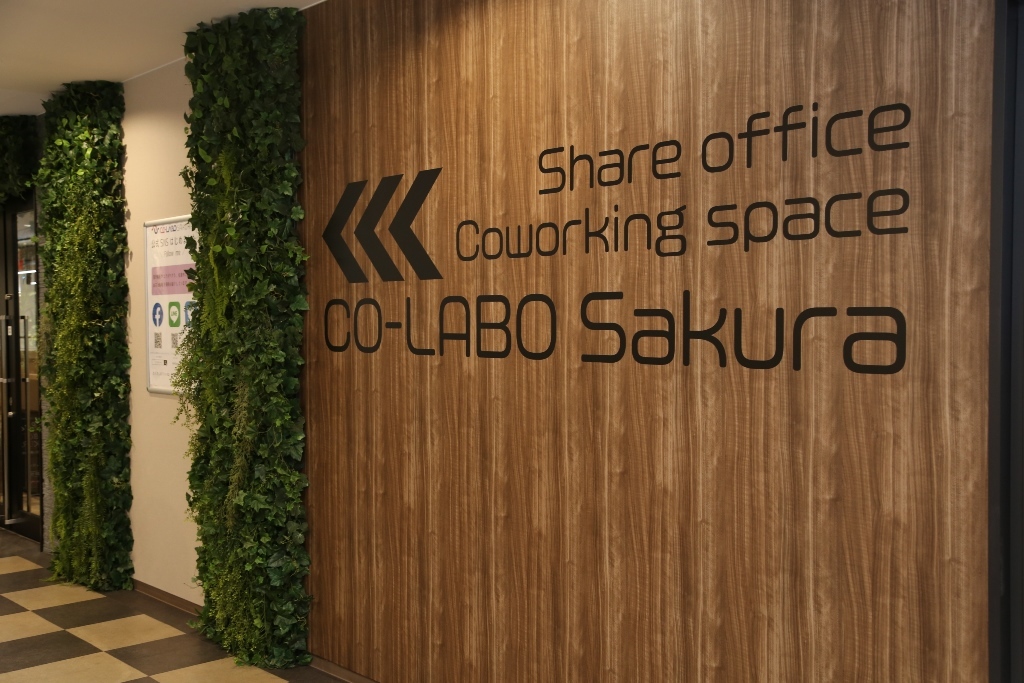木目調の壁にシェアオフィス コワーキングスペース コラボサクラと英語で書かれているコラボサクラの入口の写真