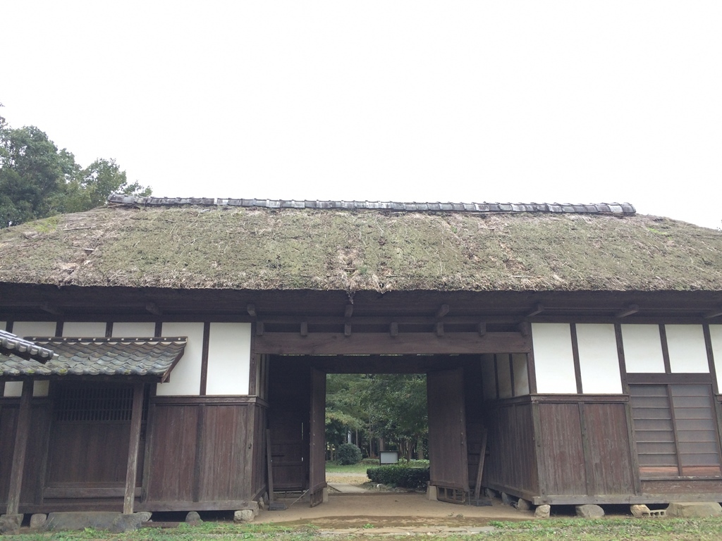 藁葺き屋根の建物の門が開いており、奥の木々が見えている写真