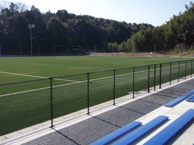 観客席の前にフェンスがあり、青いベンチから見える、白や黄色、水色の線が引かれた球技場の写真