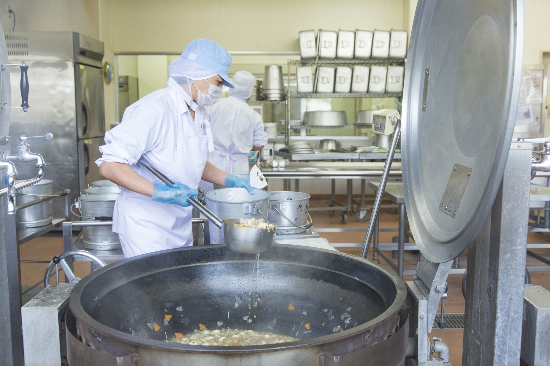 作業着姿の調理師が給食のおかずを大きな釜から食缶に移している写真