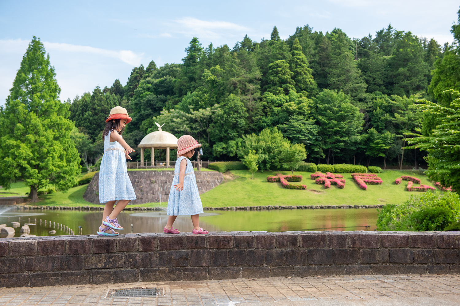 ワンピースに麦わら帽子を被った女の子2人が、池の前の石段を両手を広げて、バランスを取りながら歩いている写真