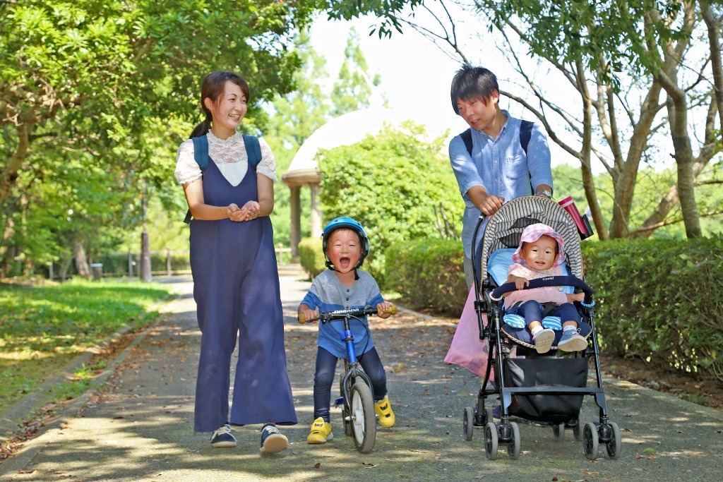 自然豊かな公園をお母さんと、自転車に乗る男の子と、女の子を乗せたベビーカーを押すお父さんが歩いている写真