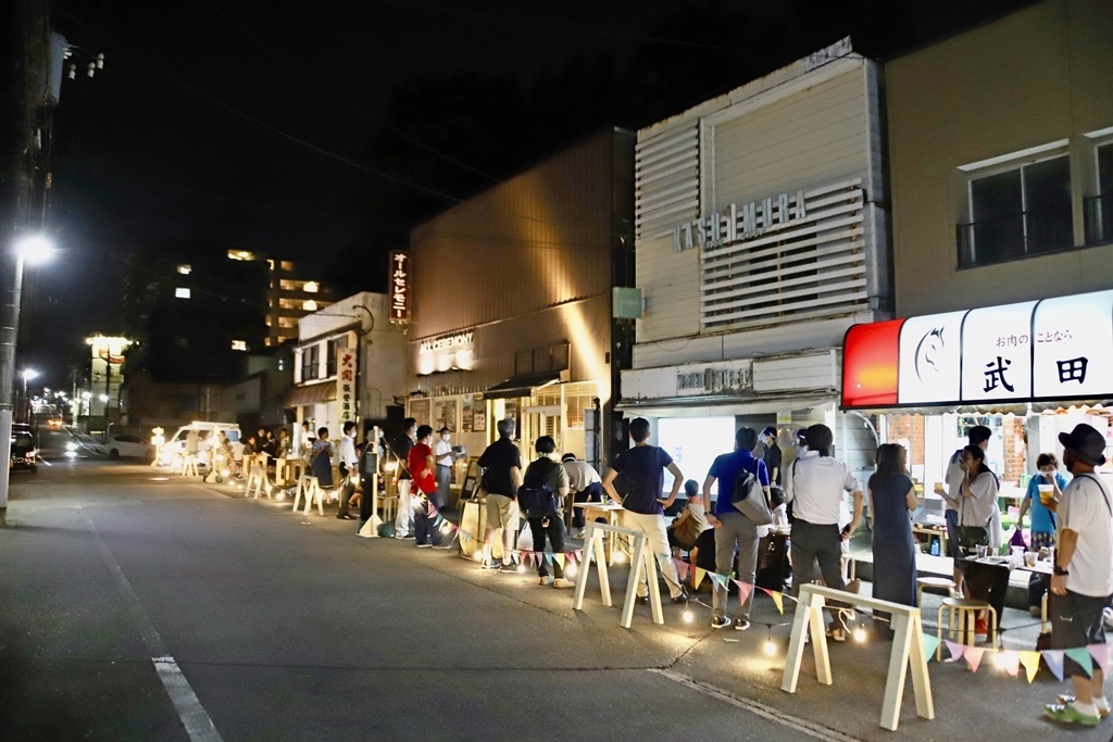 京成佐倉駅前の一方通行道路の通りの歩道と車道の間に柵を置き、ライトや飾り付けがされ、店舗前の歩道に、机やいすを置いて飲食を楽しむ方々の写真