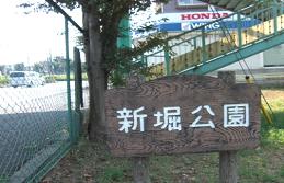 緑色のフェンスの手前に白文字で「新堀公園」と書かれた木製の看板のある新堀公園の写真