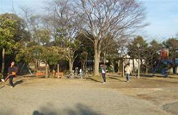 周囲に樹木が立ち並び、ベンチやすべり台が設置されている手前の広場でキャッチボールをしている親子の写真