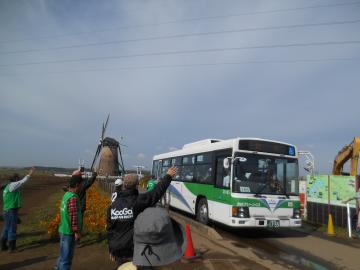 風車のある敷地内に入ってきたバスに乗っている児童に向かって手を振って出迎えているスタッフたちの写真