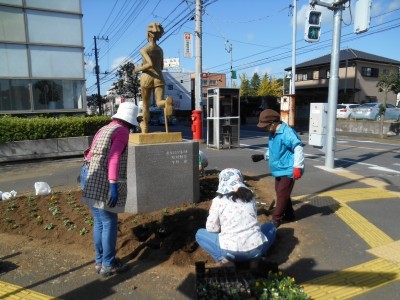 信号機の一角に設置された銅像の周囲に、3名の女性がチューリップの苗を植えている様子の写真