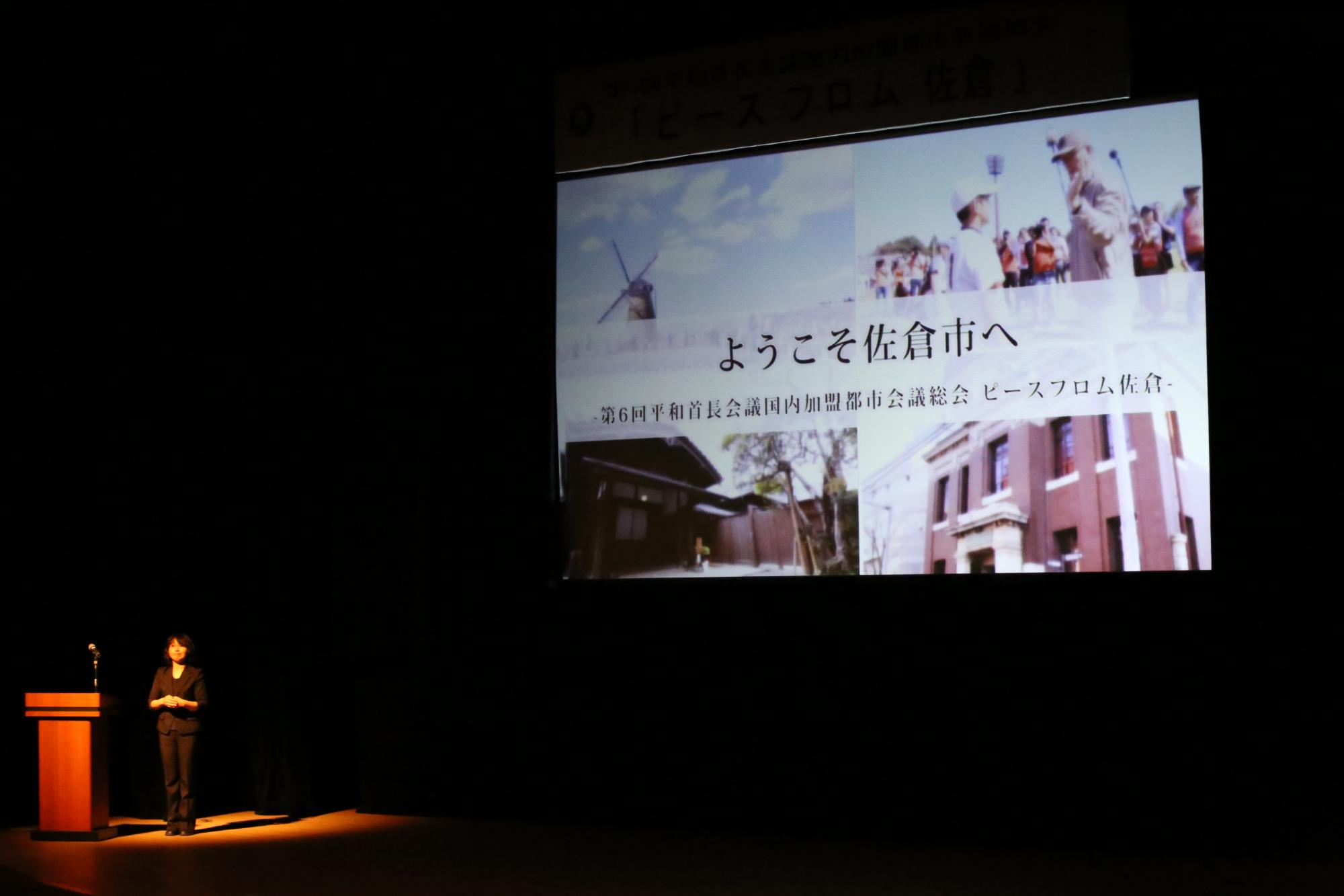 ライトアップされた演台横に立つ女性と、プロジェクトスクリーンに映る「ようこそ佐倉市へ」の映像の写真