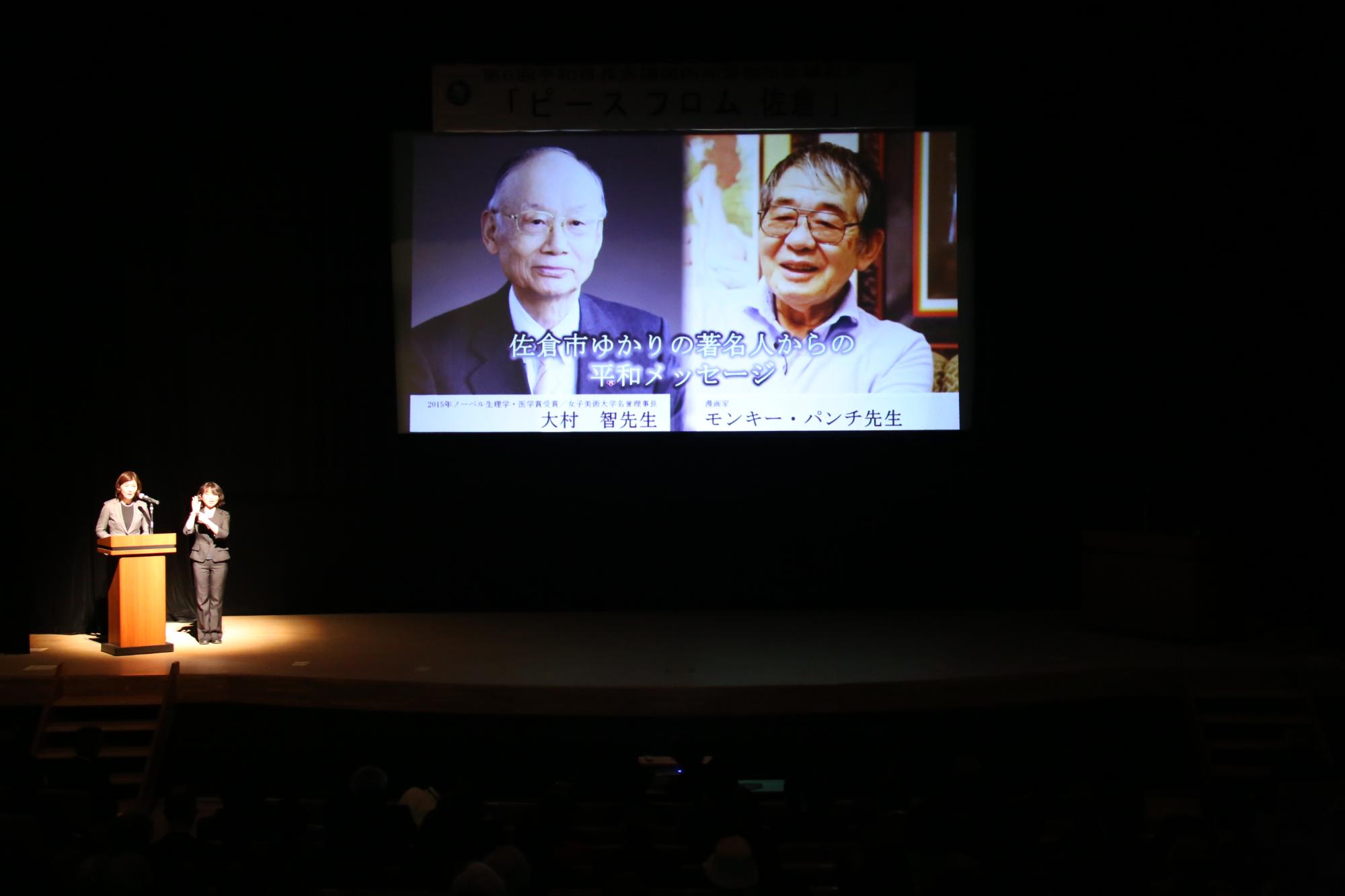 ライトアップされた演台横に立つ女性2名と、2名の著名人の写真がプロジェクトスクリーンに映っている写真