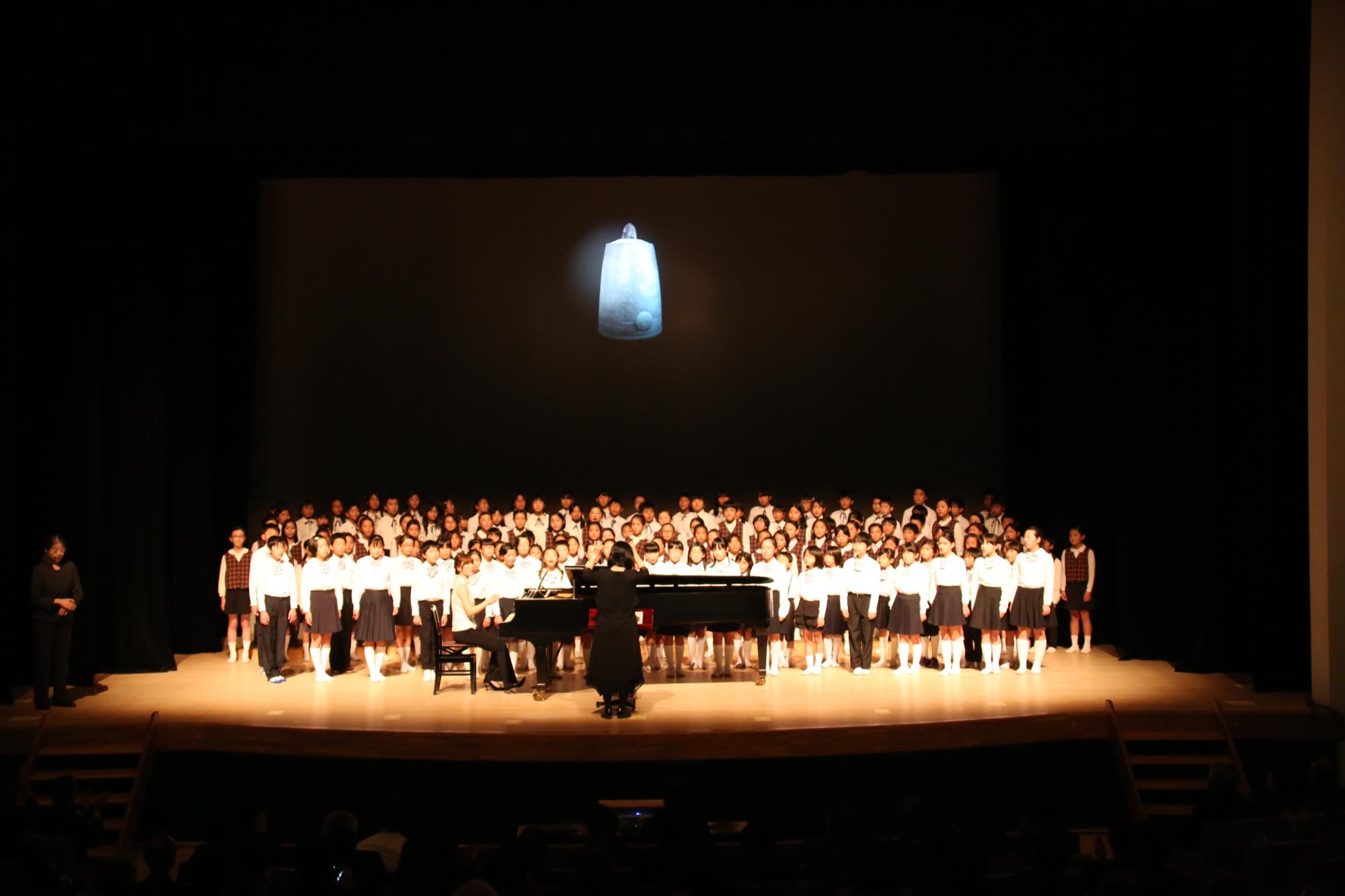 ライトアップされた舞台上の、グランドピアノの後ろに並んで合唱をしている小学校の児童の写真