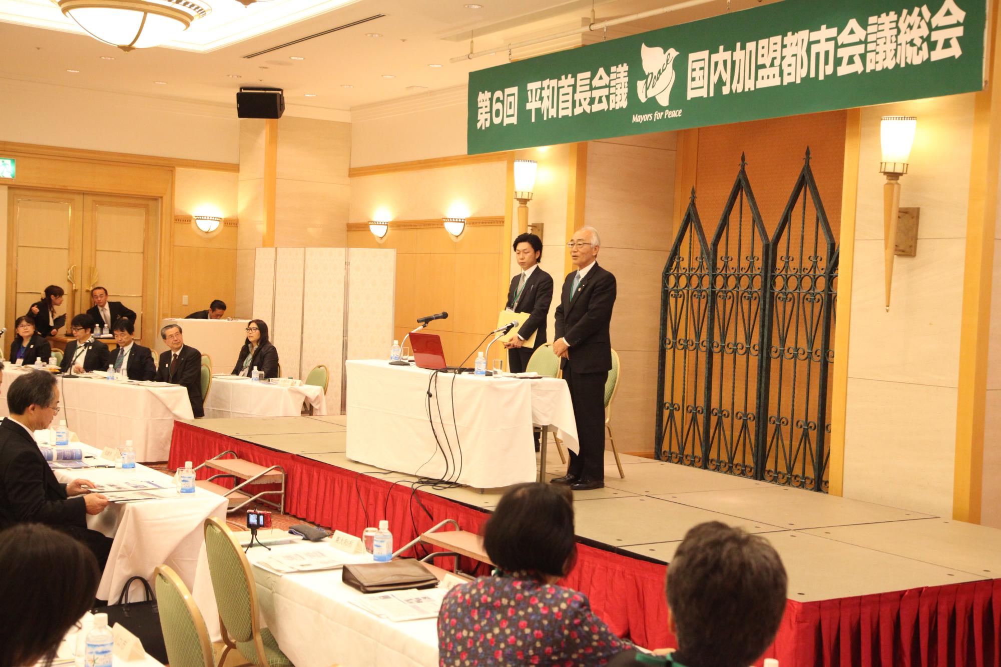 参加者が注目する中、尾崎保夫東大和市長と、男性が、議席に立っている写真