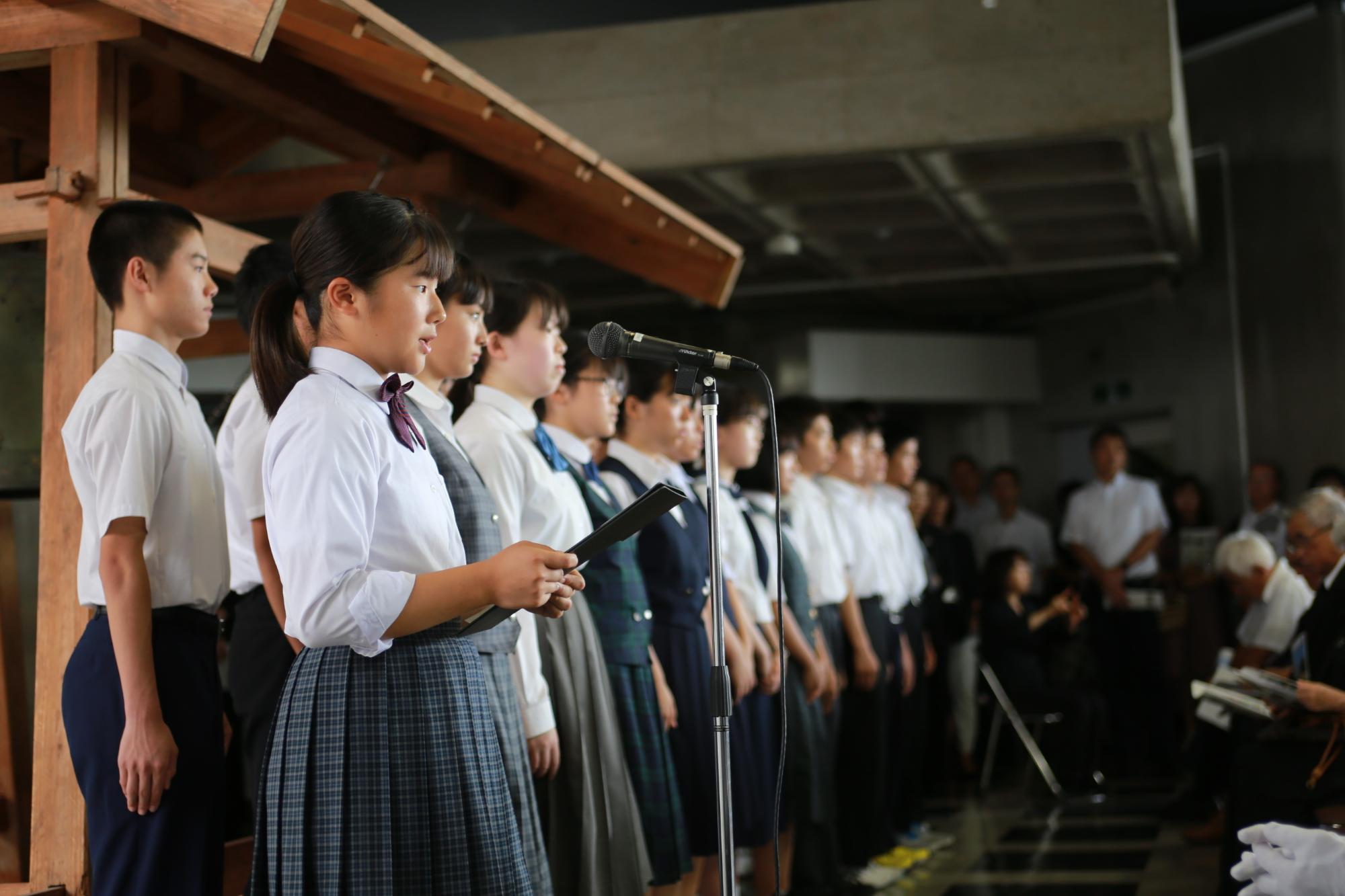 スタンドマイクの前に立つ女子学生と、並んでいる男女学生を横から写した写真