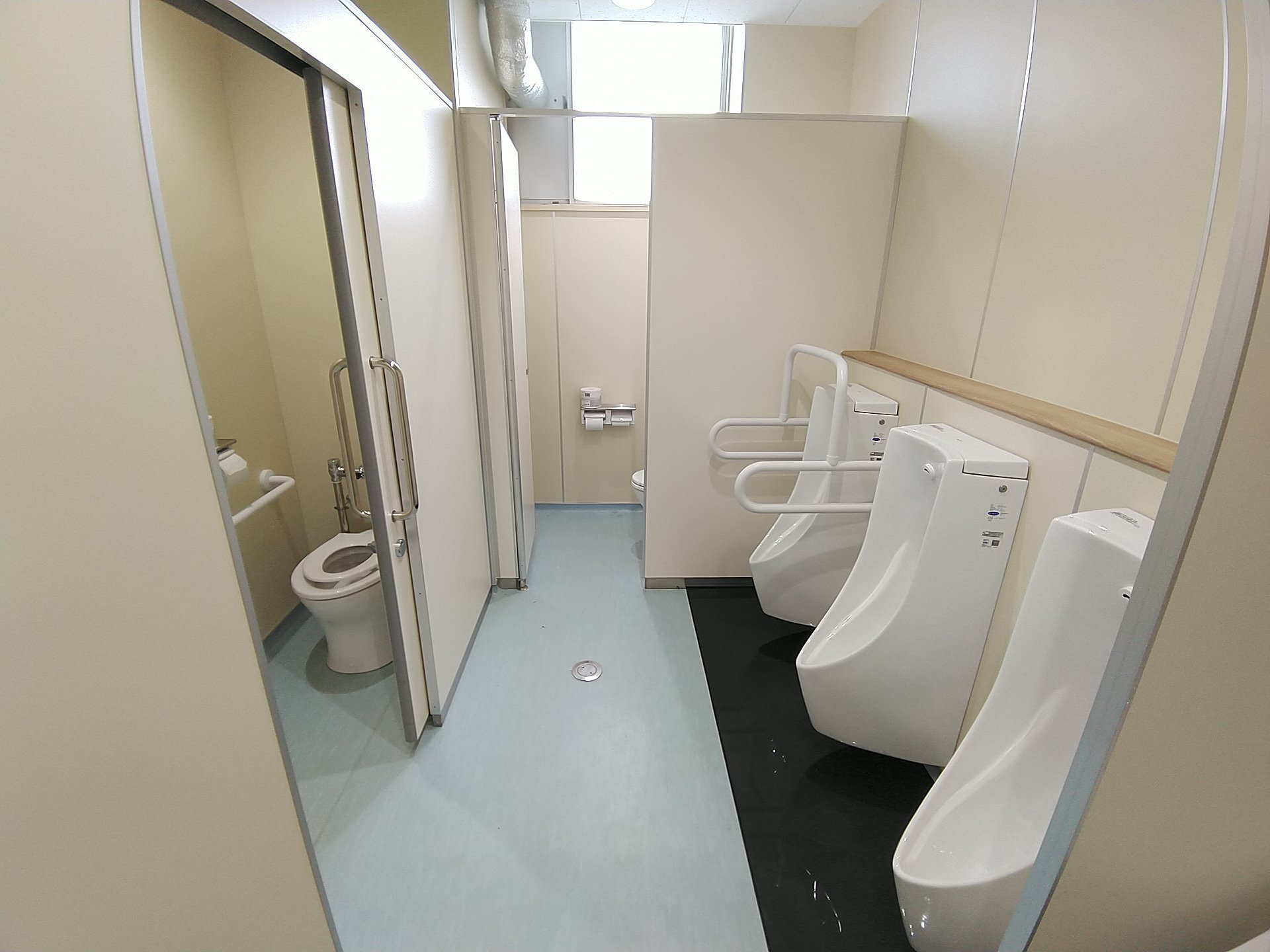 左側に洋式トイレ、右側に3つの小便器、奥のドアが開いておりトイレットペーパーホルダーがある男子トイレを入り口付近から写した写真