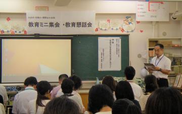 教室前の黒板右端に資料のようなものを持って立っている男性と、席に着いて話を聞いている参加者を後方から写した写真