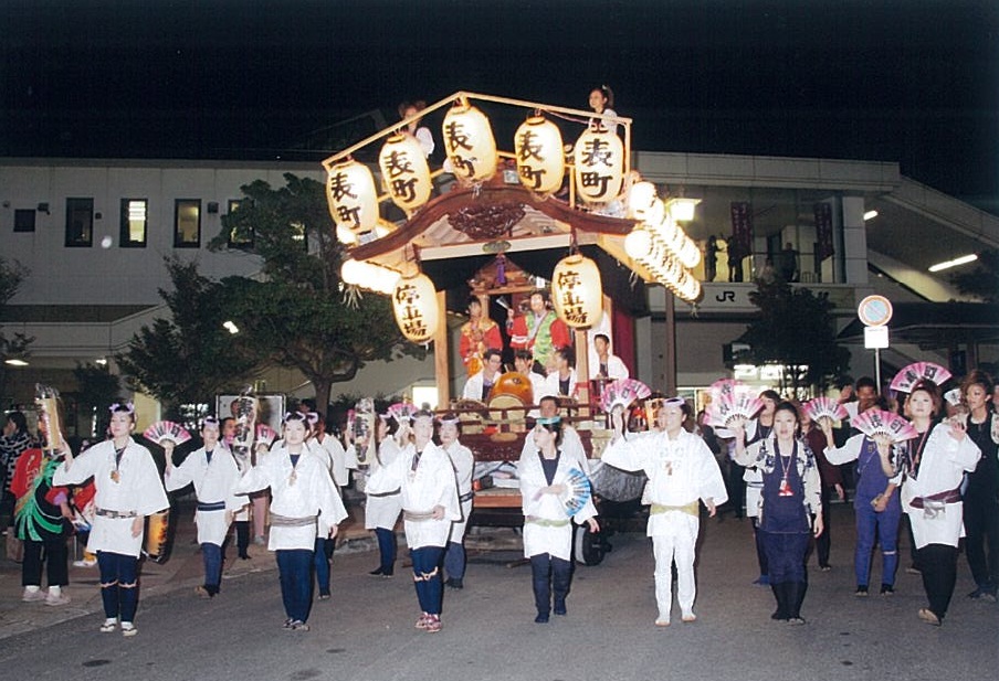 上が白い衣装を着た女性たちがハチマキをして「表町」と書かれた提灯が飾られた山車の前を歩いている写真