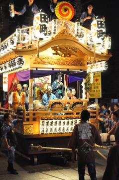 「田町」と書かれた提灯が飾られた神輿に数人の人々が乗り、夜の道路を練り歩いている写真