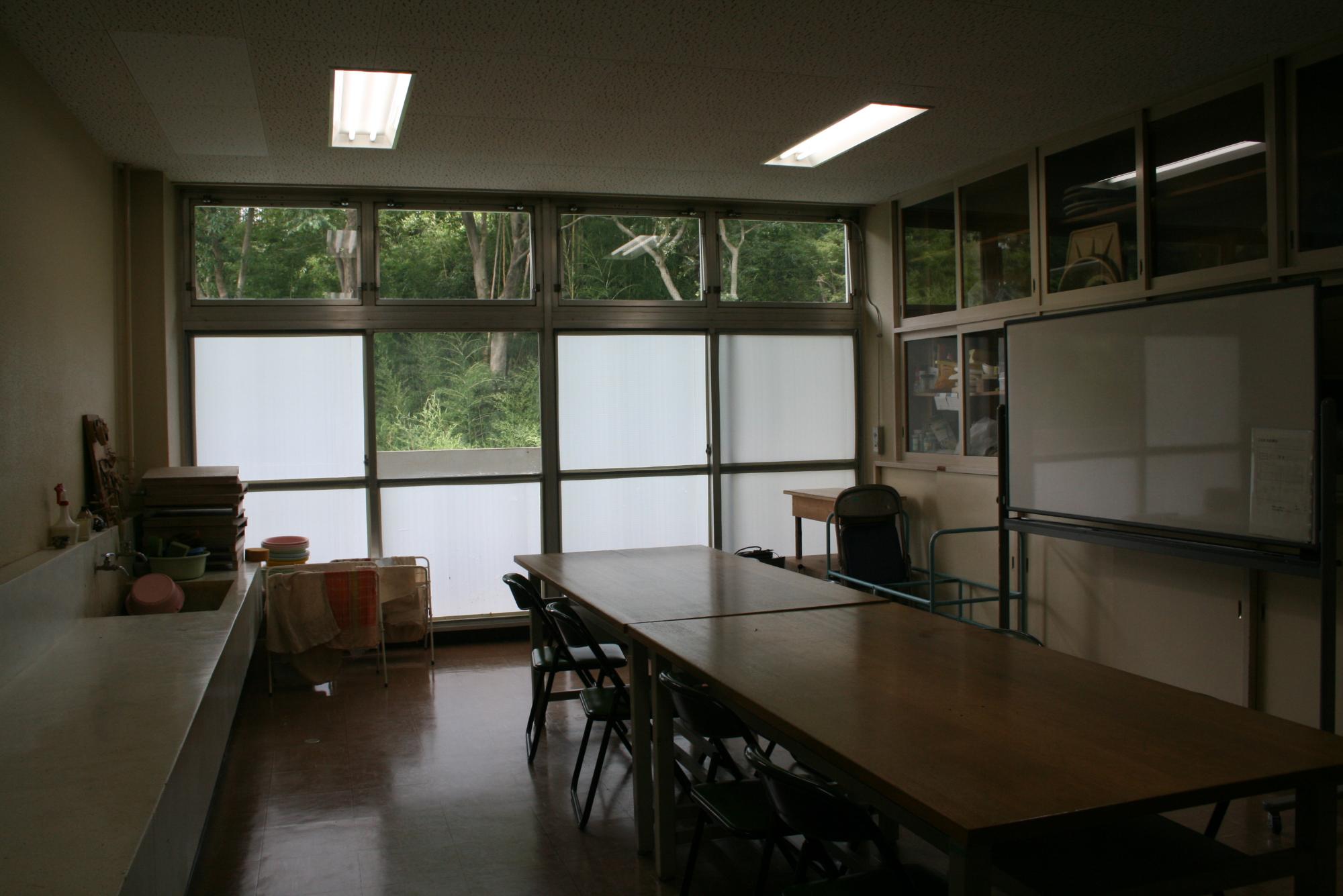 右側にホワイトボード、左側には洗い場があり、教室中央に作業台が縦に2台並び、パイプ椅子が設置されている工芸室の写真