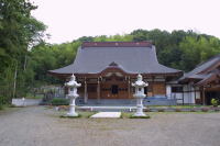 周辺を木に囲まれ、手前に2つの石碑が並ぶ大きな屋根の宝金剛寺の本堂の写真