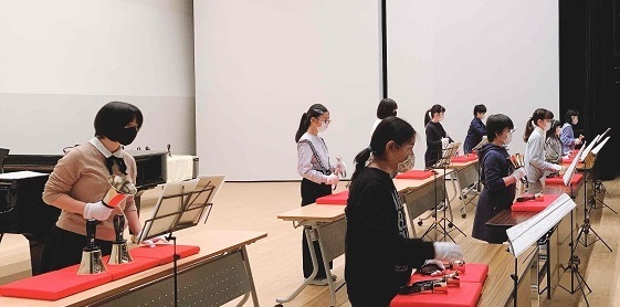舞台の上で、楽譜を見ながら演奏している少年少女ハンドベル教室の皆さんを舞台袖から写したホールでのおさらい会（2021年3月13日）の写真
