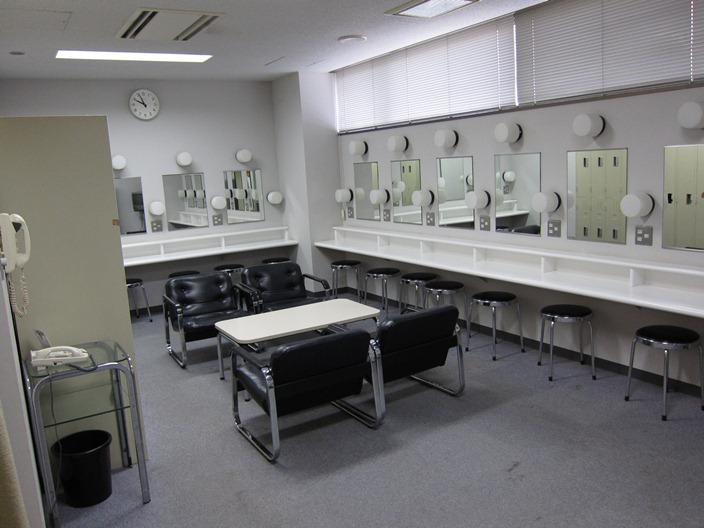 中央にテーブルと椅子が4脚設置され、壁に沿って、メイク用の鏡と丸椅子が並んでいる楽屋1の写真