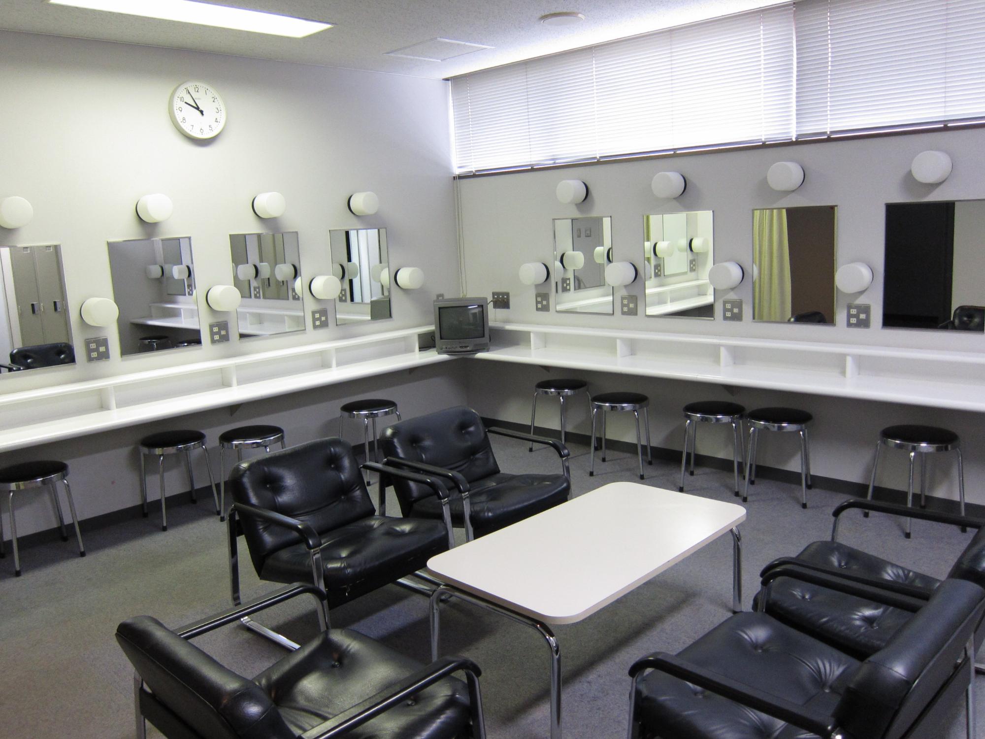 中央にテーブルと椅子が5脚設置され、壁にはメイク用の鏡と丸椅子が並んでいる楽屋2の写真