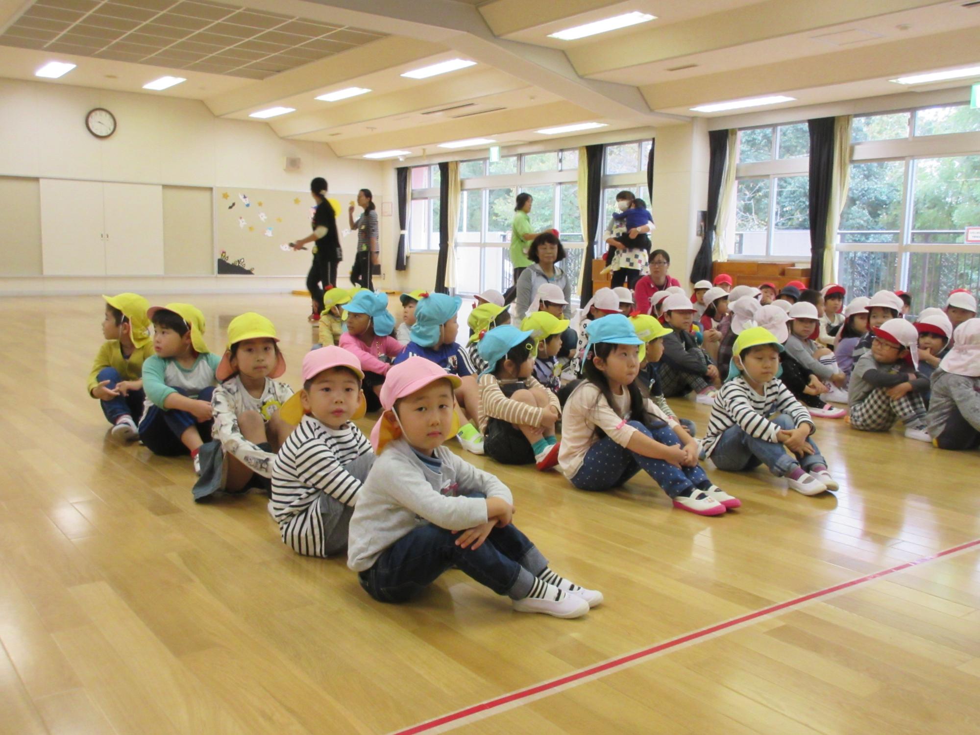 園児たちがホールの床に縦並びに体育座りをしている写真