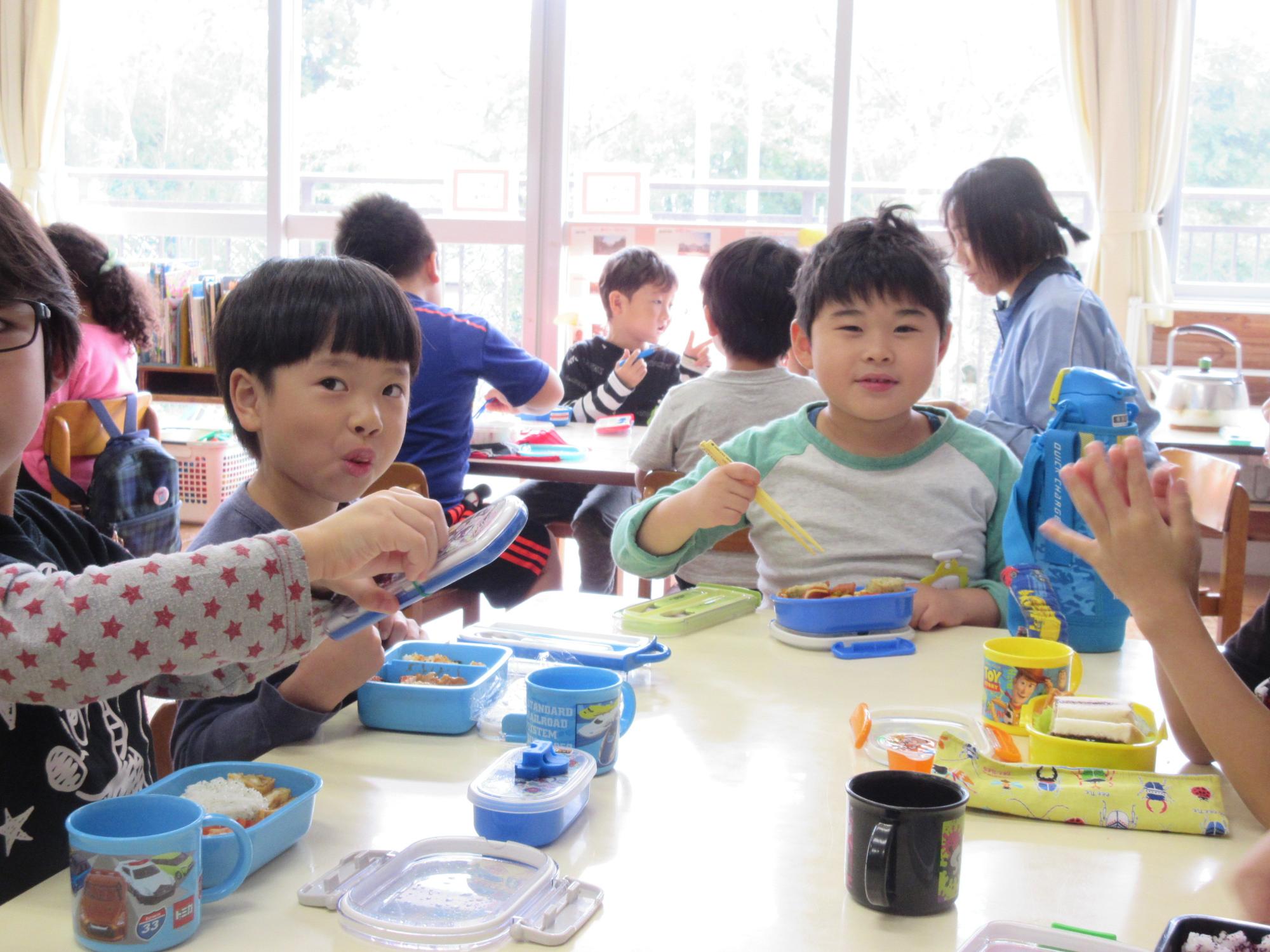 園児たちがグループに分かれて座り、机の上に置いたお弁当を食べている写真