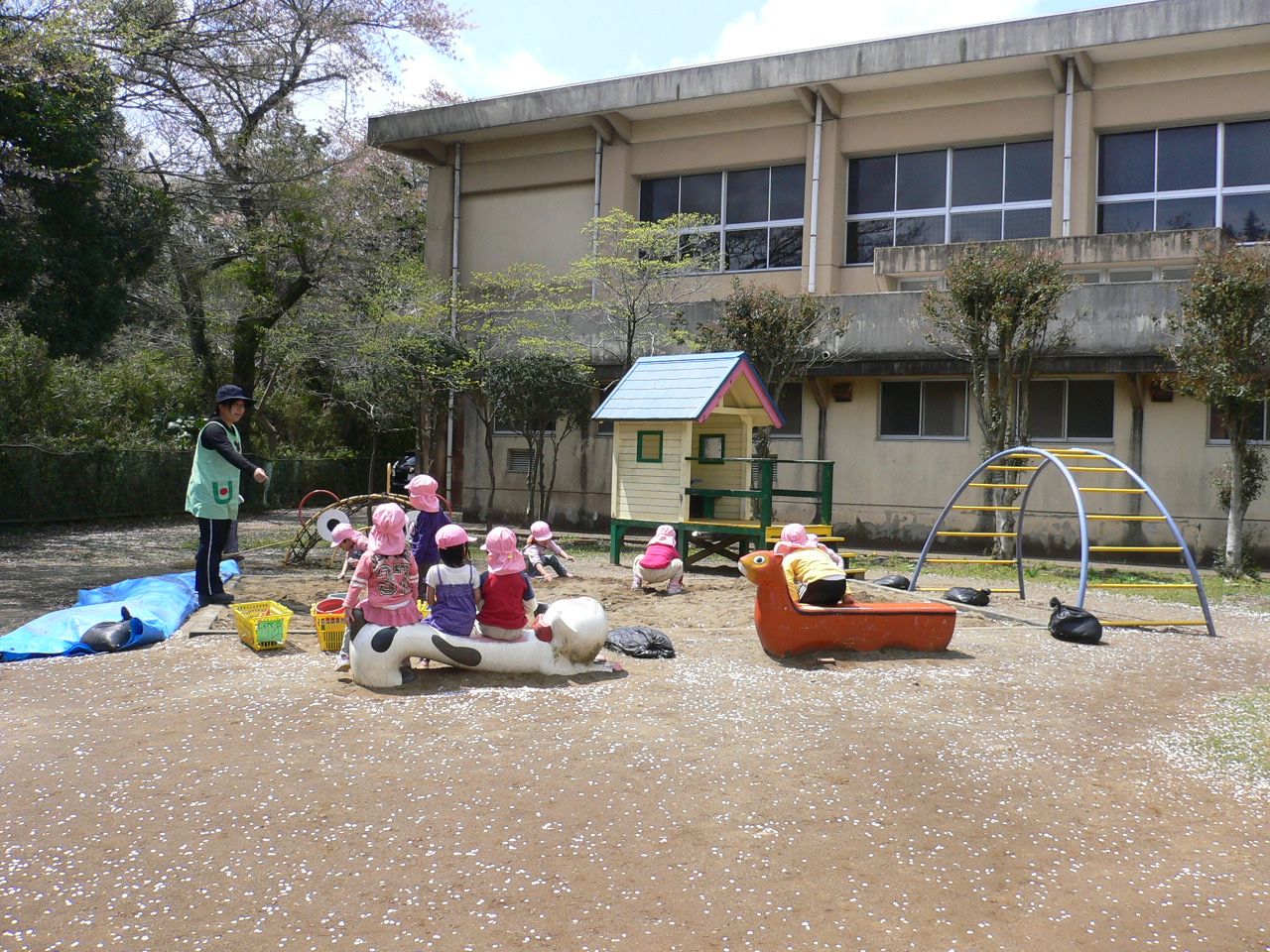 園庭で園児や先生が、遊具に座ったり砂場で遊んだりしている写真