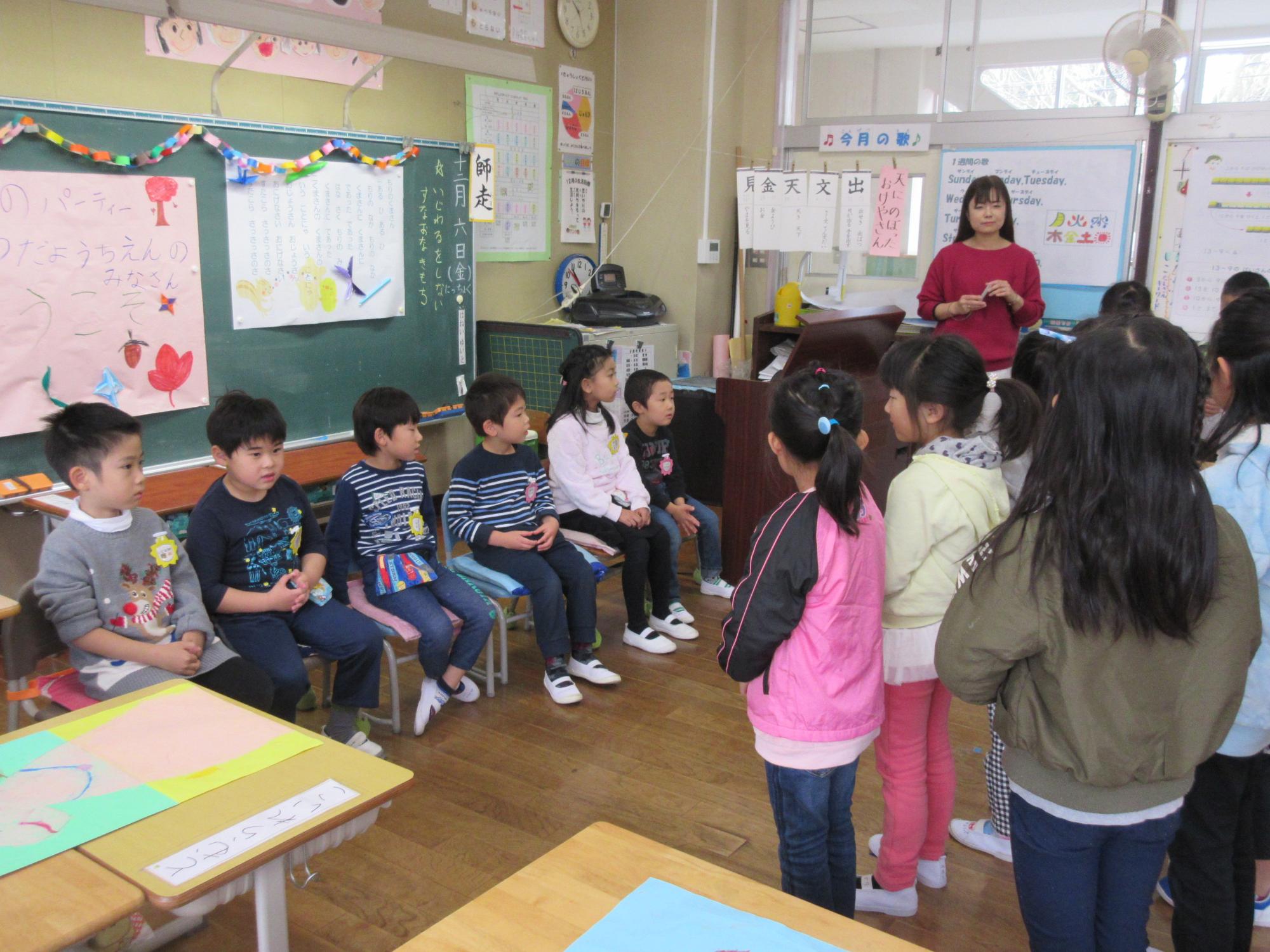 小学校の教室で、黒板の前に着席している園児と、向かい合って立っている小学生の児童の写真