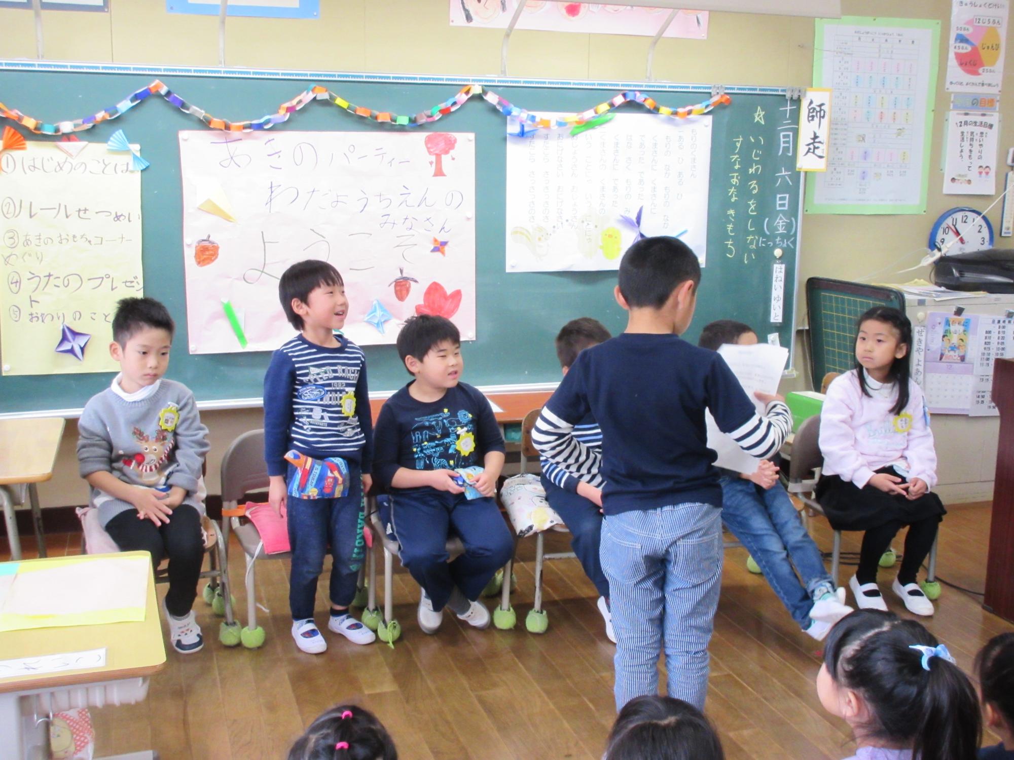 向かい合って着席している園児と児童の間に、紙を持った男の子が立っている写真