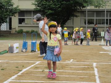 校庭に白線をひいた横断歩道を、帽子を被った女の子が右手を挙げて渡っている写真