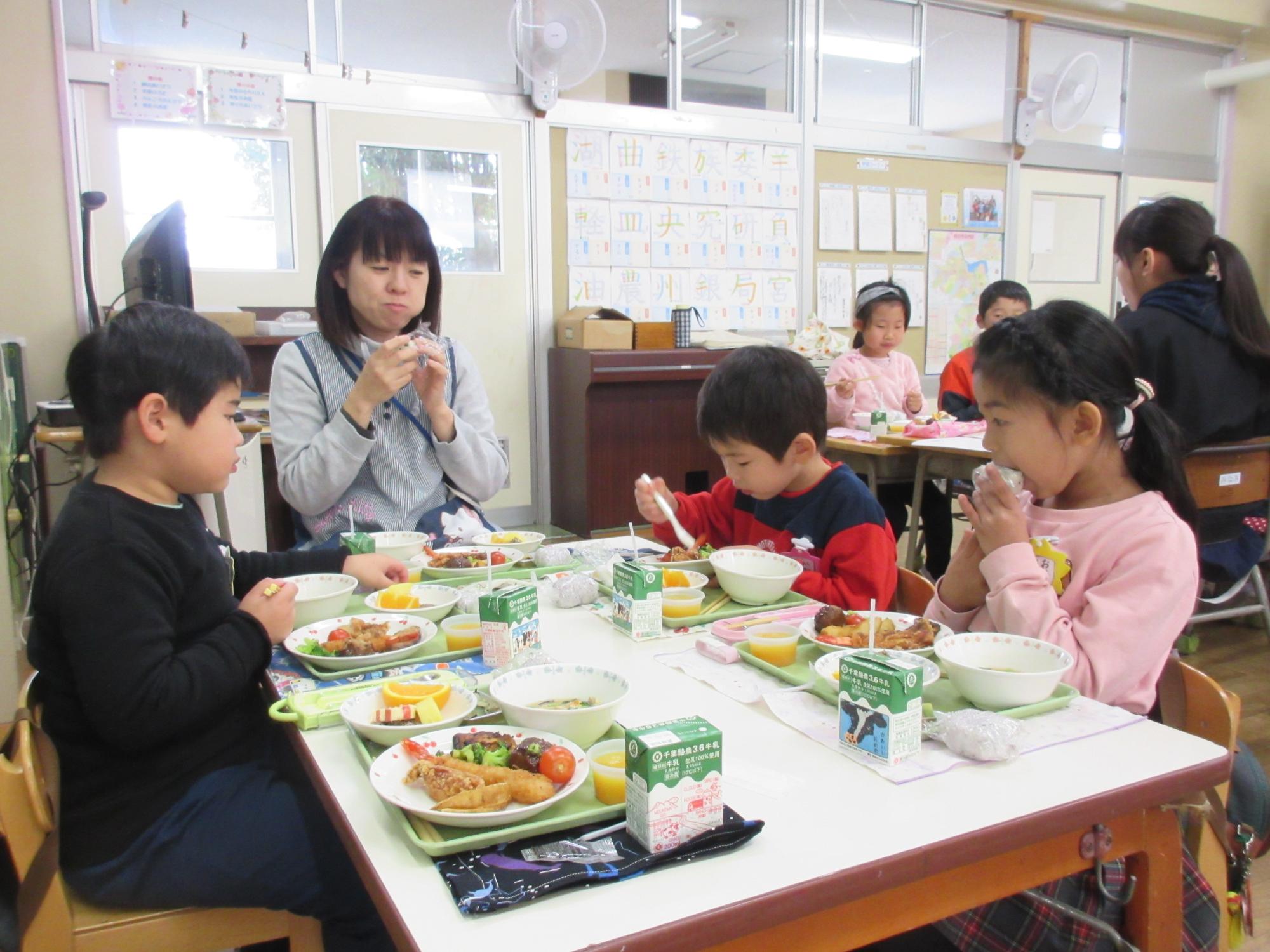 園児3名と女性が、トレイに並べられたお皿に盛りつけられた給食を食べている写真