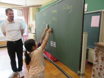 ALTの先生が笑顔で見ている前に設置された黒板に2名の子どもが立ち右手を挙げている、クイズに挑戦している様子の写真