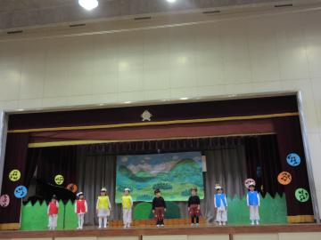 赤や黄色、黒、青の衣装を着た園児たちが舞台上に横並びに並んでいる写真