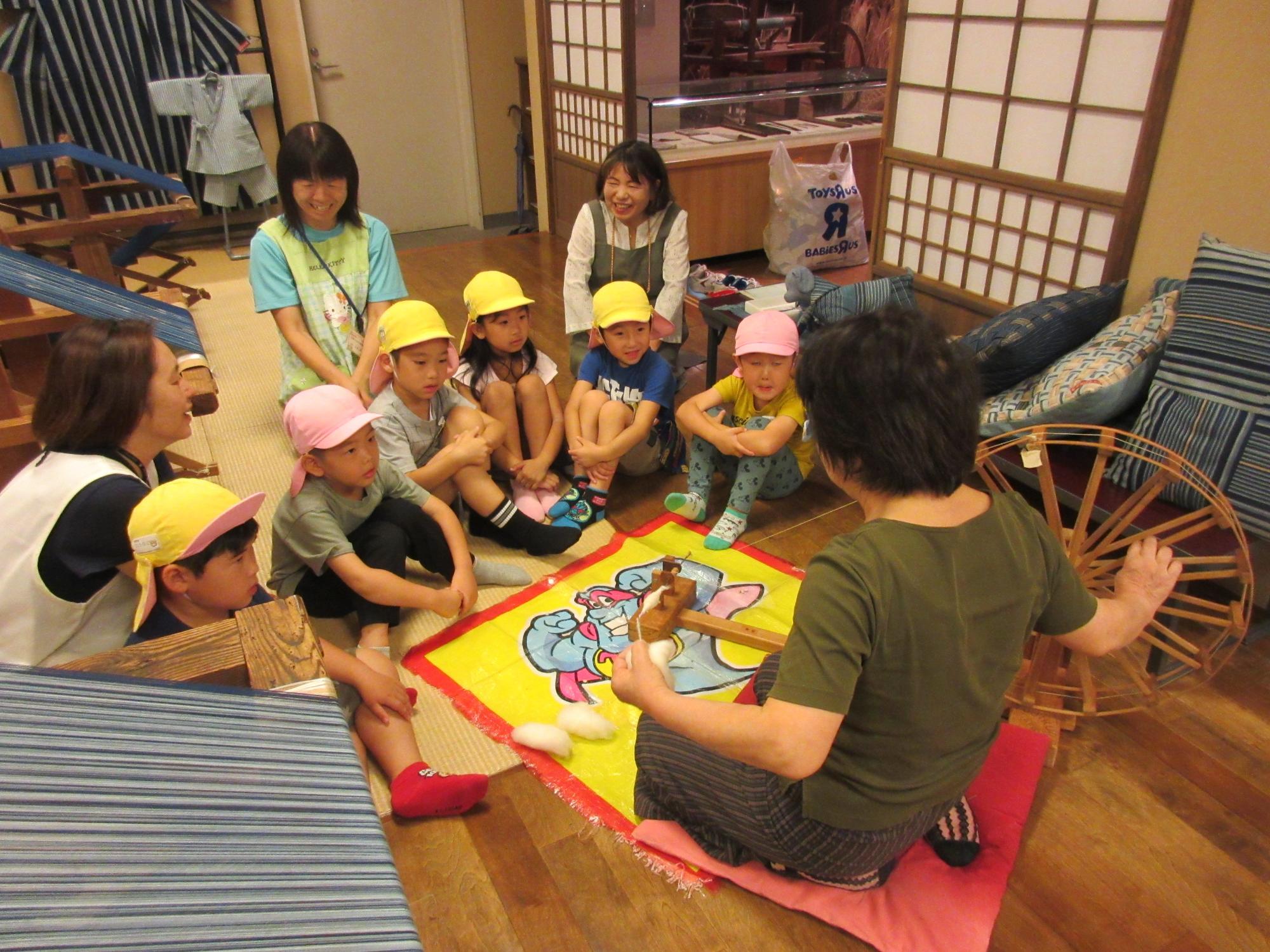 綿を紡いで糸にする道具の周りに園児たちが集まり、座って説明を聞いている写真