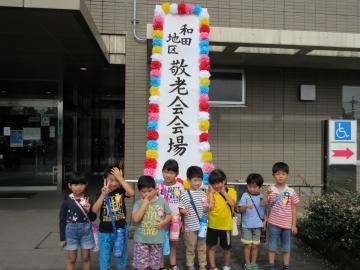 紙花が飾られた敬老会会場と書かれた看板の前で、水筒を肩から掛けた園児8名が並んでいる写真