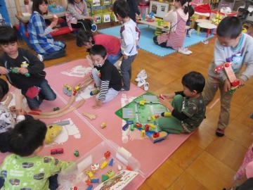 ピンクのマットの上に園児たちが座り、小さな乗り物のおもちゃで遊んでいる写真