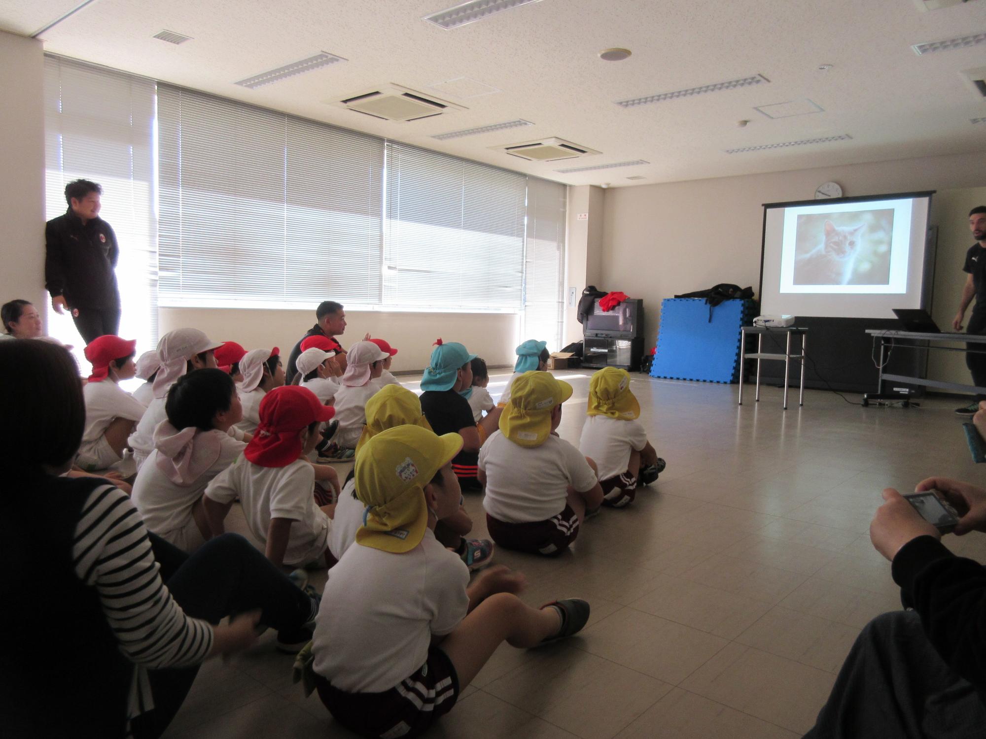 体操服で帽子を被った園児が並んで座り、正面のプロジェクタースクリーンに映る映像を見ている写真