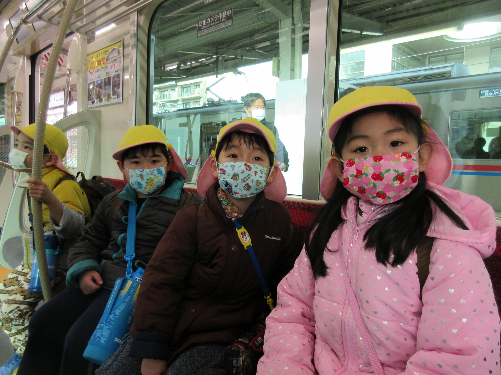 黄色い帽子にマスクをつけた園児たちが電車の座席に座っている写真
