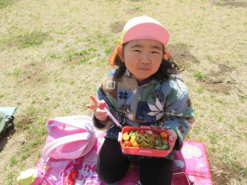 一人の女の子の園児が片手にお弁当をもって、反対の手にお箸を持ちながらピースをしている写真