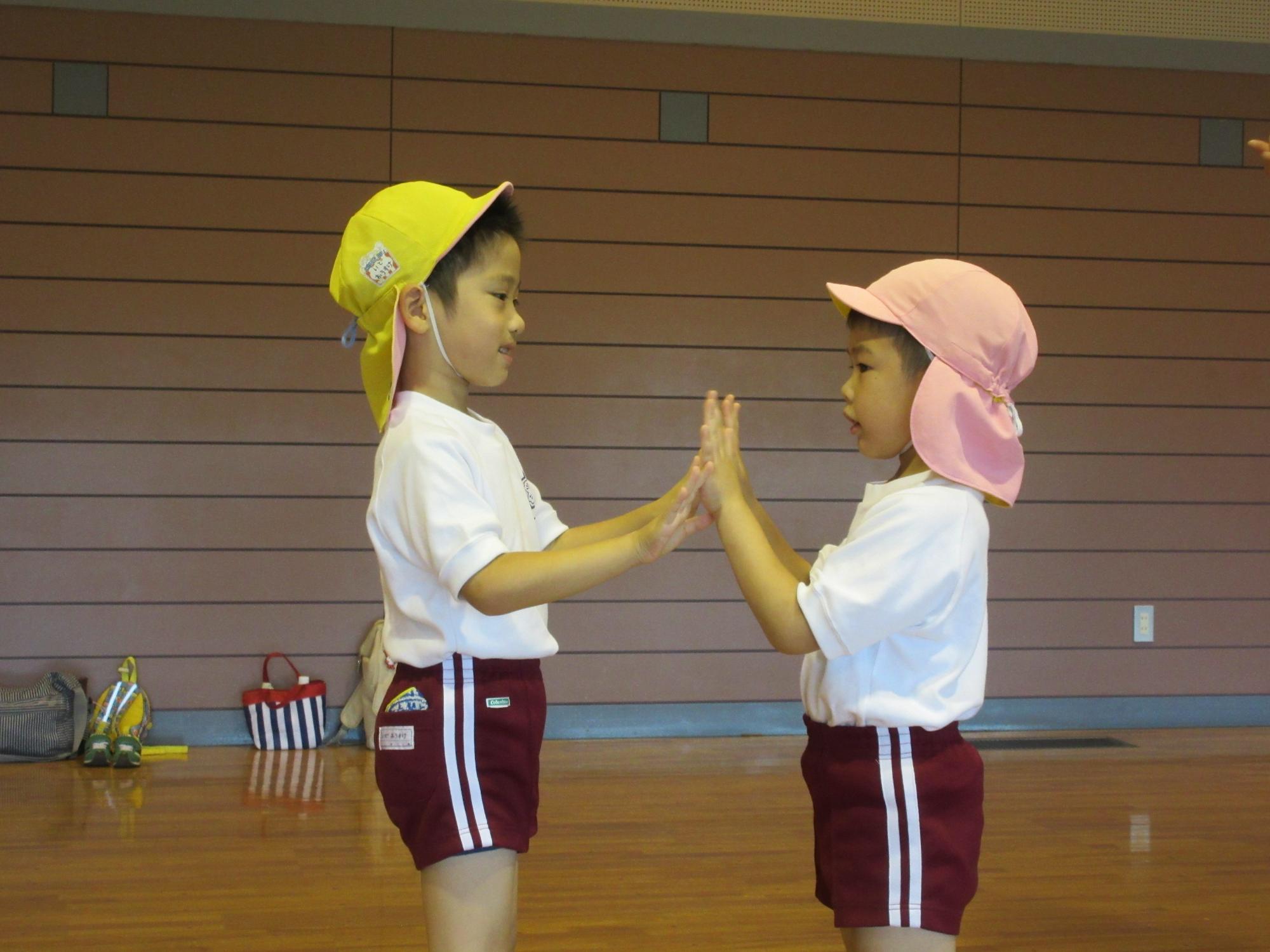 ピンクの帽子と黄色の帽子を被った園児2名が向かい合って両手を合わせている写真
