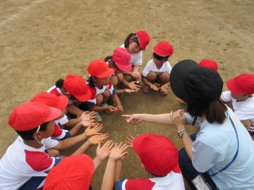 赤い帽子を被った園児や小学生、職員が輪になって座り、手のひらを上に向けて出している写真