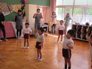 体操着を着た年少組の子ども達が間隔をあけて、右手を挙げて体操しており、教室の端に立っている保護者の方々が写真を撮ったり子ども達の様子を参観している様子の写真