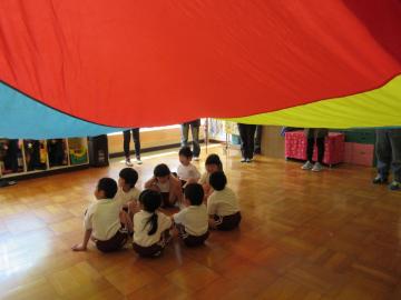 教室の上空でカラフルな色の大きなパラバルーンが膨らみ、中央に集まって、座っている子供たちの写真