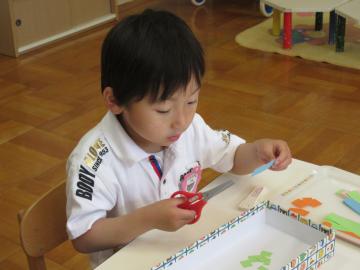 椅子に座っている男の子が、はさみを使って水色の紙を切ろうとしている写真