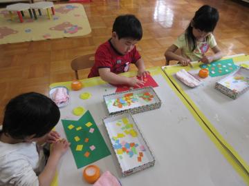3名の子ども達が椅子に座り、糊を使って色画用紙に小さく切った紙のウロコを貼っている写真