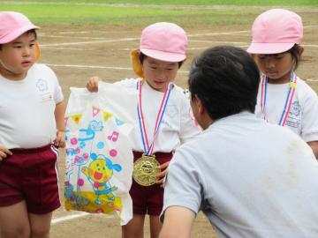 ピンクのカラーキャップを被った園児が、メダルを付けてプレゼントをもち、園長先生の前に立っている写真