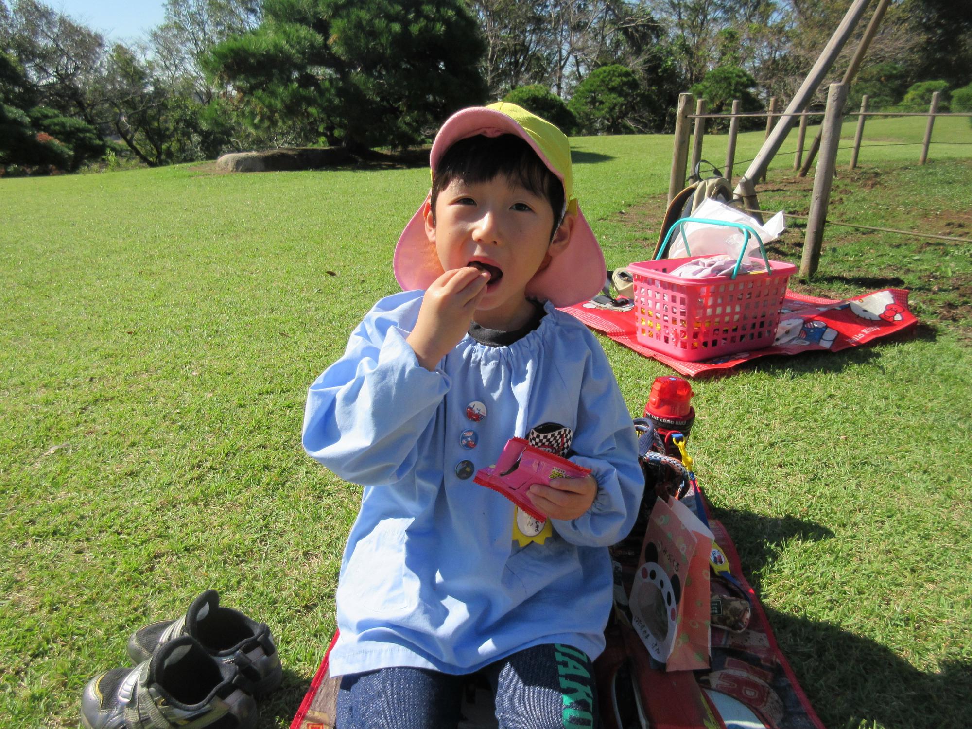 水色のスモックを着た園児が片手にお菓子の袋を持っておやつを口に入れている写真
