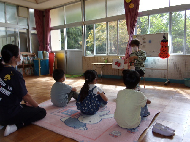 司書の先生が、3人の園児と女性の前で絵本の読み聞かせをしている写真